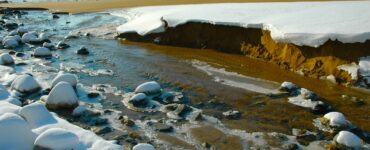 A river mouth at the beach of a larger body of water, with broken snowbanks along the river's edge and ice attached to rocks in the river.