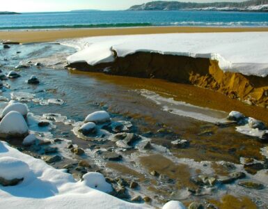 A river mouth at the beach of a larger body of water, with broken snowbanks along the river's edge and ice attached to rocks in the river.