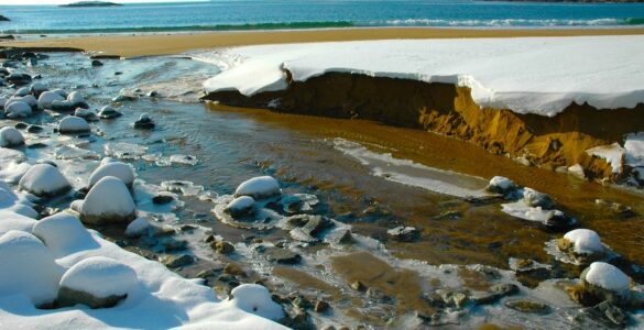 A river mouth at the beach of a larger body of water, with broken snowbanks along the river's edge and ice attached to rocks in the river.