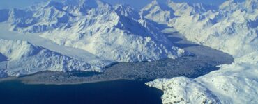 A wide view of glaciers with frozen flows flowing into a bay. The sky is clear and blue, and snow-covered mountains surround the scene. Ice floats in the water.