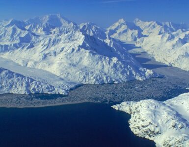 A wide view of glaciers with frozen flows flowing into a bay. The sky is clear and blue, and snow-covered mountains surround the scene. Ice floats in the water.