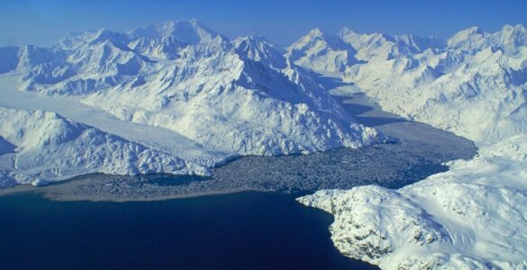 A wide view of glaciers with frozen flows flowing into a bay. The sky is clear and blue, and snow-covered mountains surround the scene. Ice floats in the water.