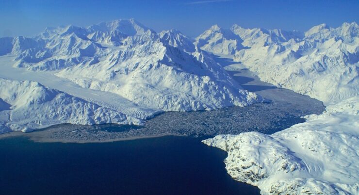 A wide view of glaciers with frozen flows flowing into a bay. The sky is clear and blue, and snow-covered mountains surround the scene. Ice floats in the water.