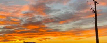 A sunset with vibrant, colorful clouds behind a mountain range. In the foreground, power lines are visible, indicating the presence of the aging infrastructure of a small desert community.