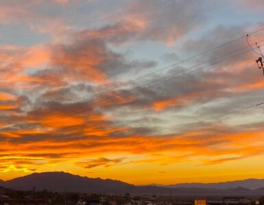 A sunset with vibrant, colorful clouds behind a mountain range. In the foreground, power lines are visible, indicating the presence of the aging infrastructure of a small desert community.