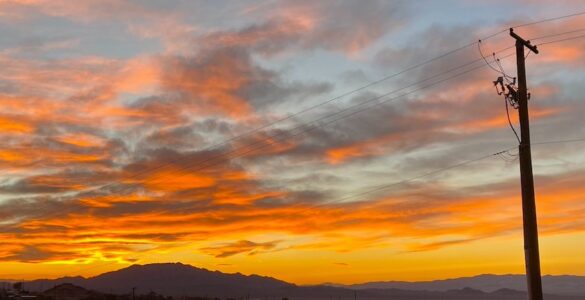 A sunset with vibrant, colorful clouds behind a mountain range. In the foreground, power lines are visible, indicating the presence of the aging infrastructure of a small desert community.