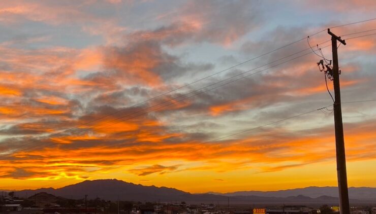 A sunset with vibrant, colorful clouds behind a mountain range. In the foreground, power lines are visible, indicating the presence of the aging infrastructure of a small desert community.