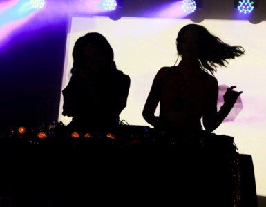 Silhouettes of two DJs performing at a music event with colorful stage lighting.