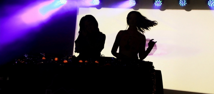 Silhouettes of two DJs performing at a music event with colorful stage lighting.