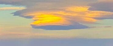 A lenticular cloud shaped like a saucer, with orange and blue hues. The mountains below are faintly visible in the fading light of dusk.