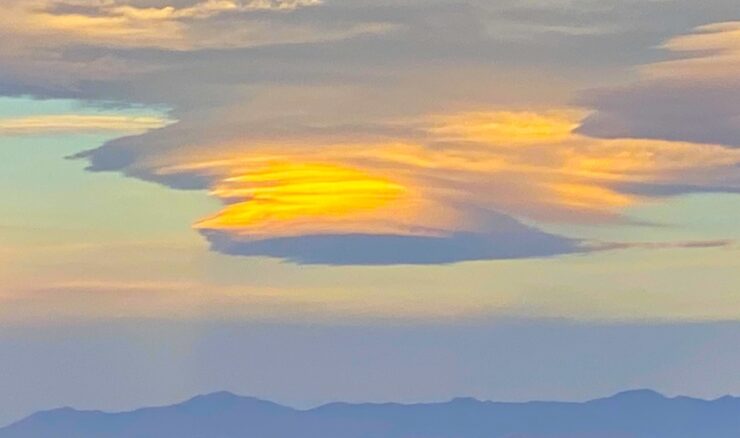 A lenticular cloud shaped like a saucer, with orange and blue hues. The mountains below are faintly visible in the fading light of dusk.